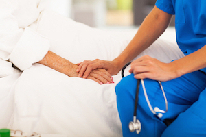 Female Nurse is sitting on the edge of a swing bed and holding an elderly patients hand
